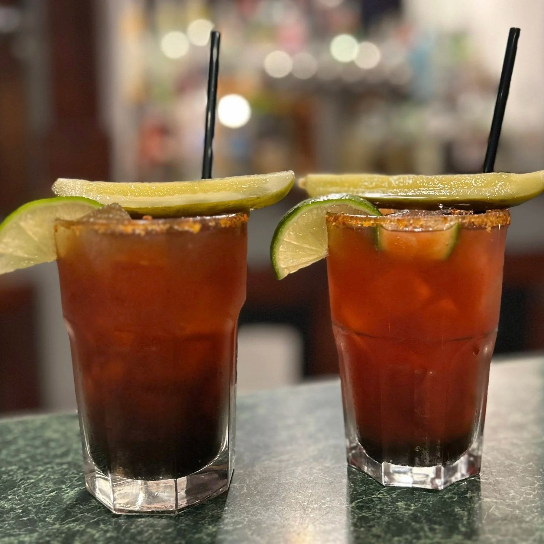 Two cocktails in tall glasses garnished with lime slices and salted rims, with black straws, on a bar counter.