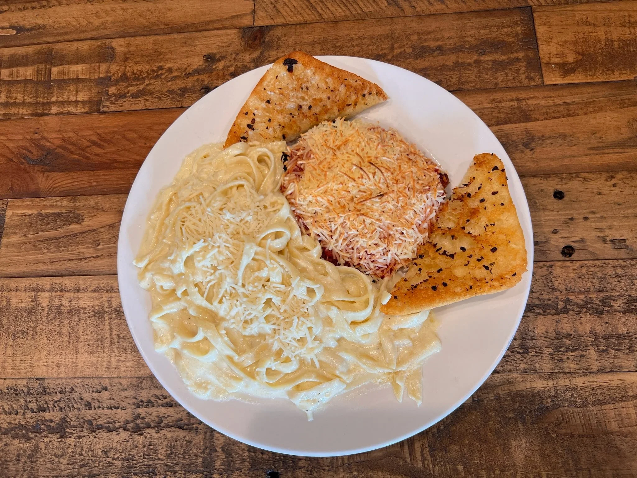 Fettuccine Alfredo with shredded cheese, two pieces of garlic bread, and a serving of tuna salad, on a white plate on a wooden table.
