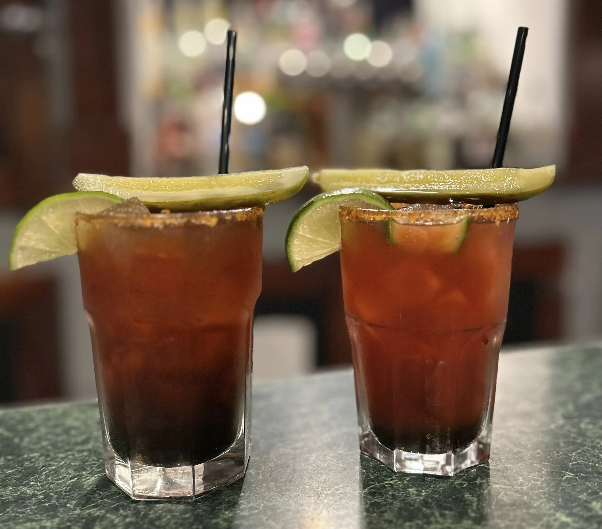 Two tall glasses of dark red cocktail garnished with lime and lemon wedges, with black straws, on a dark countertop in a bar or restaurant setting.