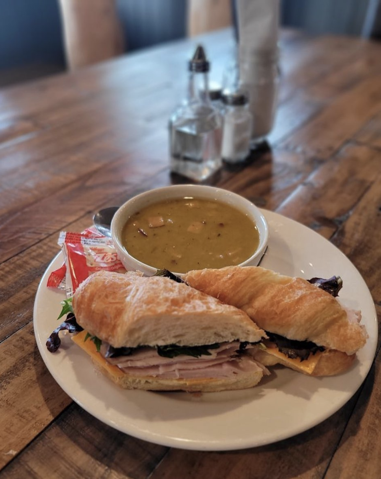 Eddy's Bar and Grill serving a plate with a croissant sandwich containing ham, cheese, lettuce, and tomato, served with a bowl of soup and two small containers of salt and pepper on a wooden table.