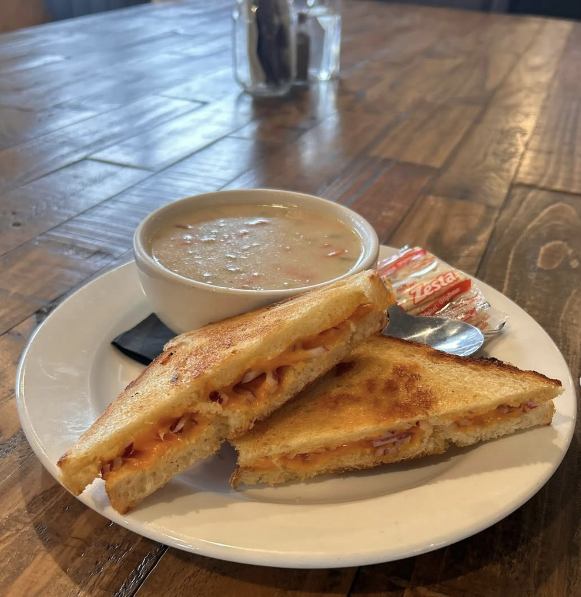 Eddy's Bar and Grill serving a plate with grilled cheese sandwiches, a bowl of soup, a packet of crackers, and a spoon on a wooden table.
