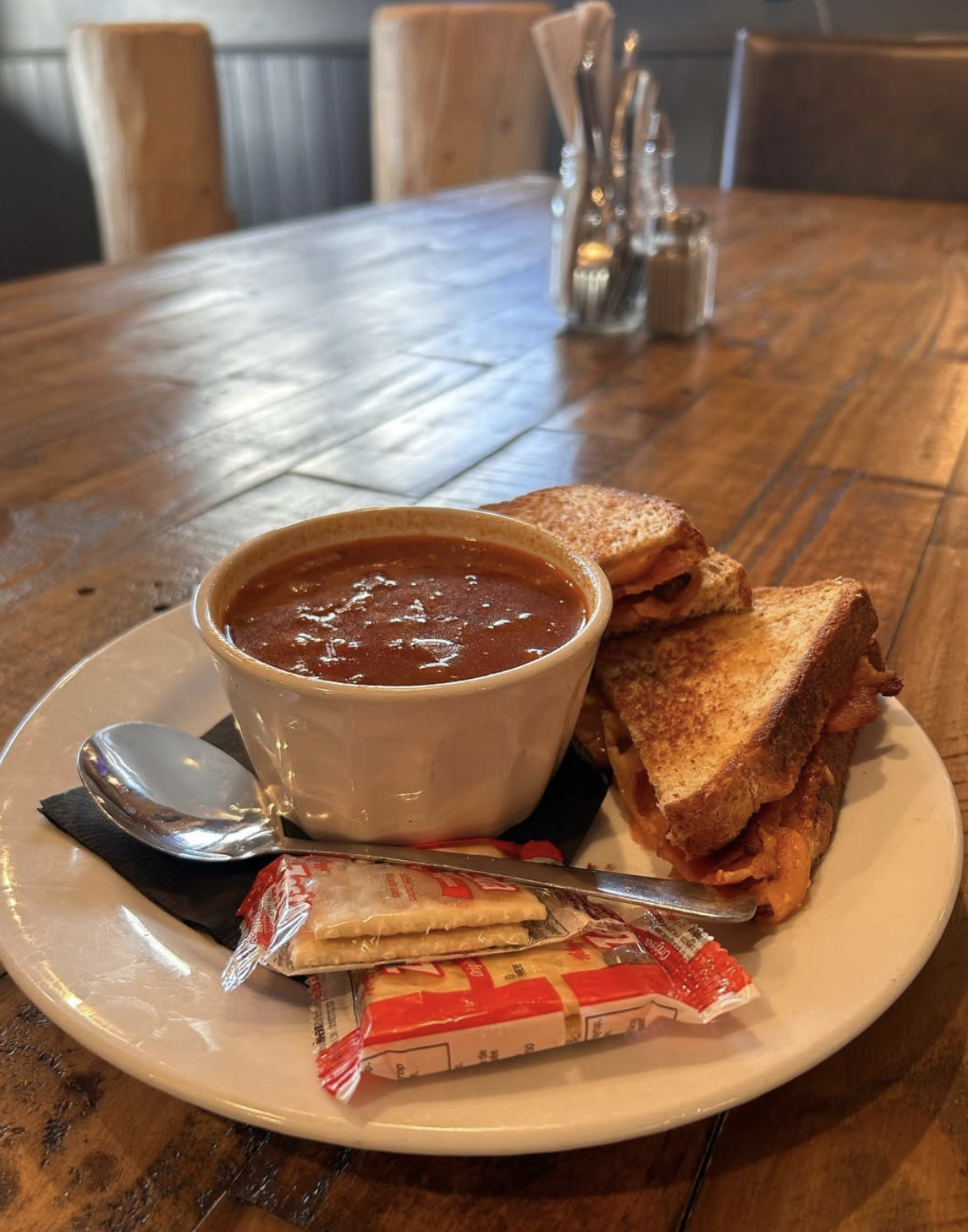 A plate with a bowl of chili, two grilled cheese sandwiches, a spoon, a packet of saltines, and a disposable spoon on a wooden table in a restaurant setting.
