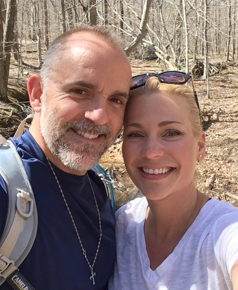 A smiling man and woman taking a selfie outdoors in a wooded area during the day.