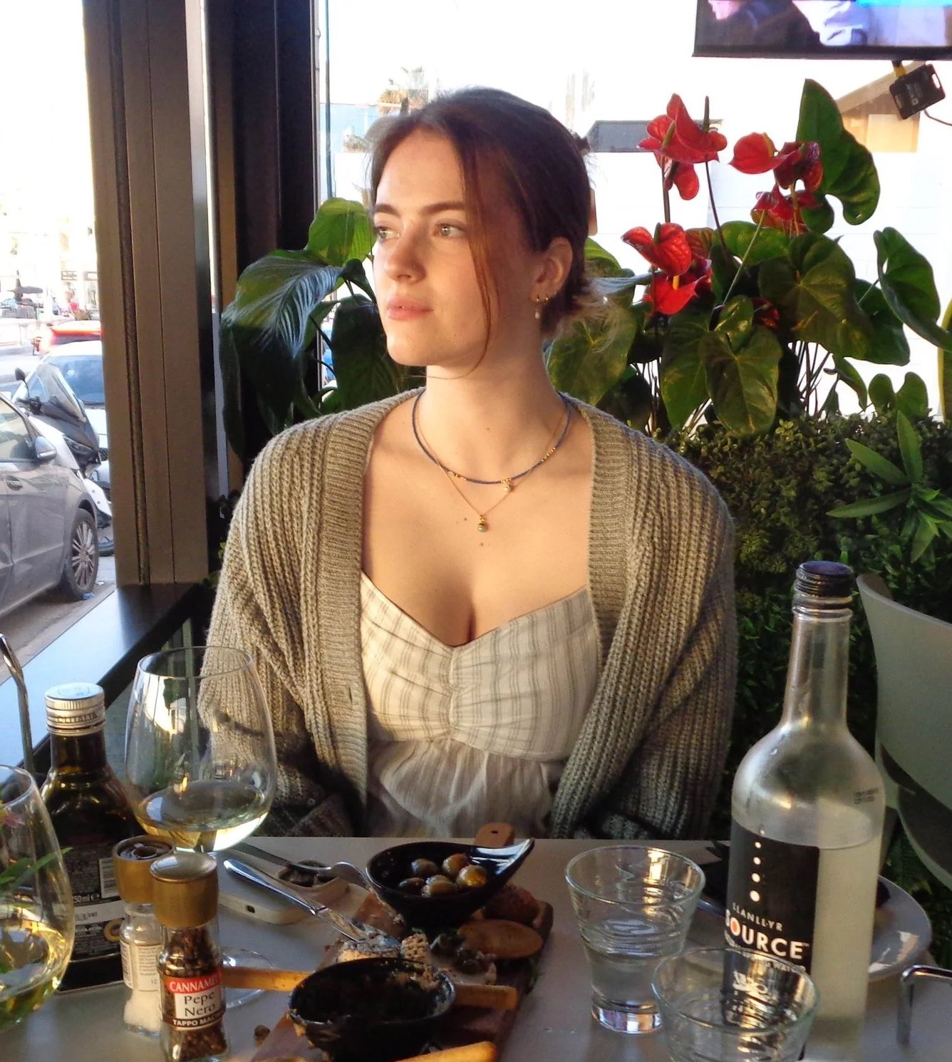 A woman with short brown hair and light skin seated at a restaurant table near a window, surrounded by green plants and red flowers, with food and drinks on the table.