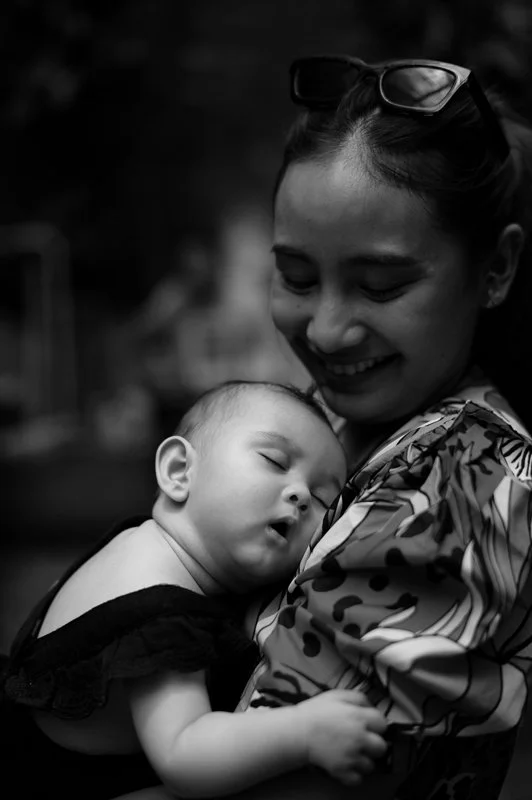 a mom smiling lovingly at her baby while they sleep on her chest