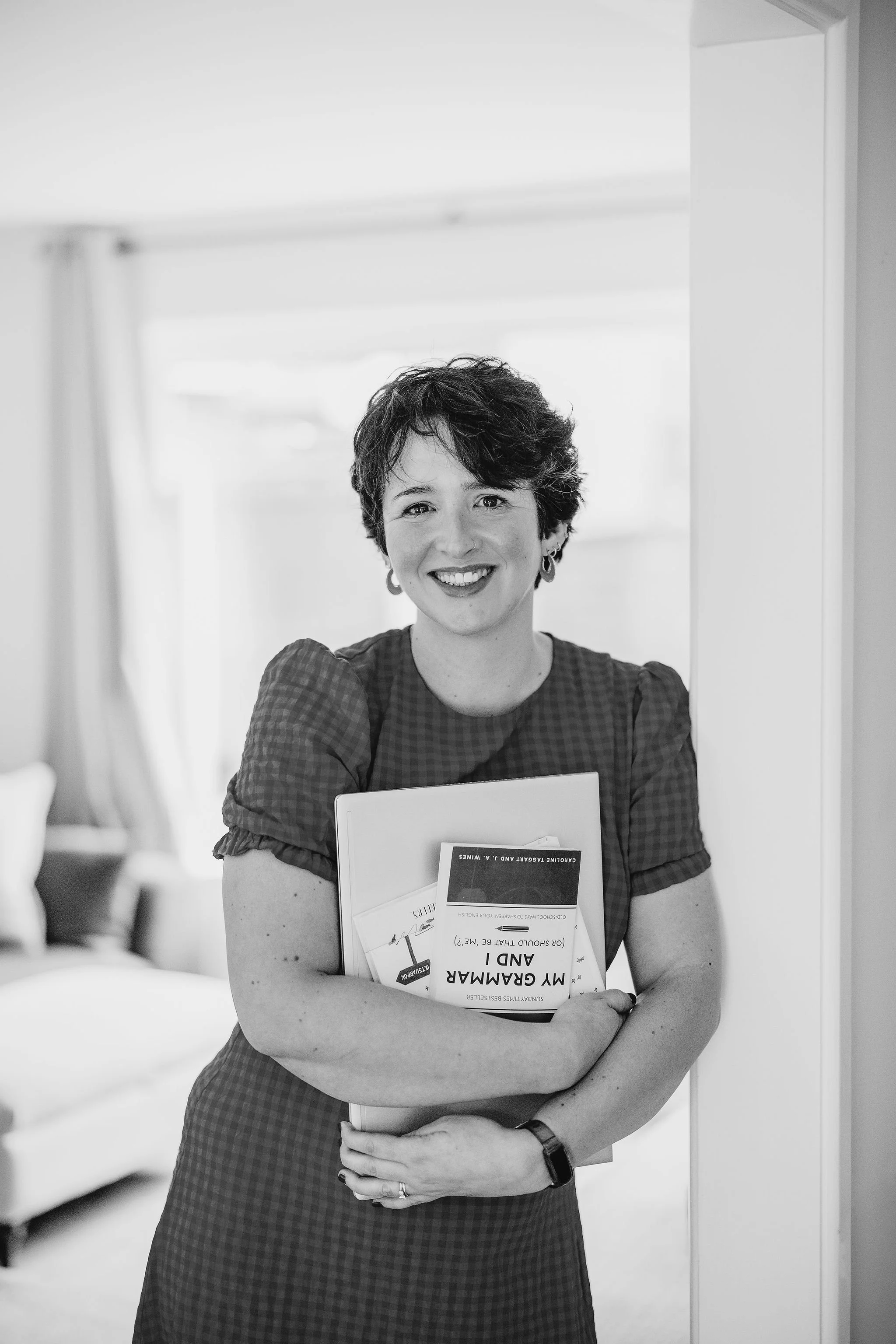 A woman with short, curly hair and earrings smiling while holding a book and a folder in a room with soft lighting.