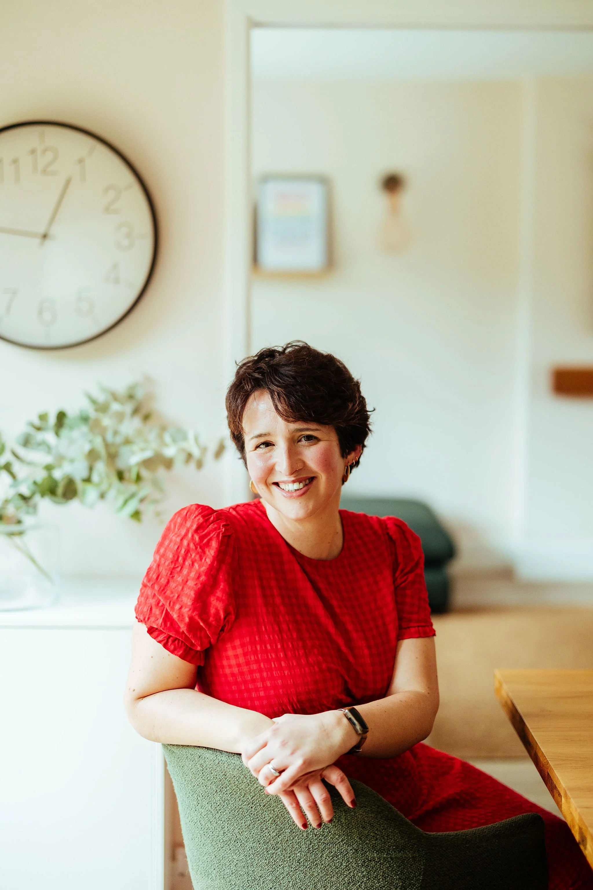 A woman with short dark hair, wearing a red dress, smiling and seated in a green chair in a bright room with flowers and a clock on the wall.