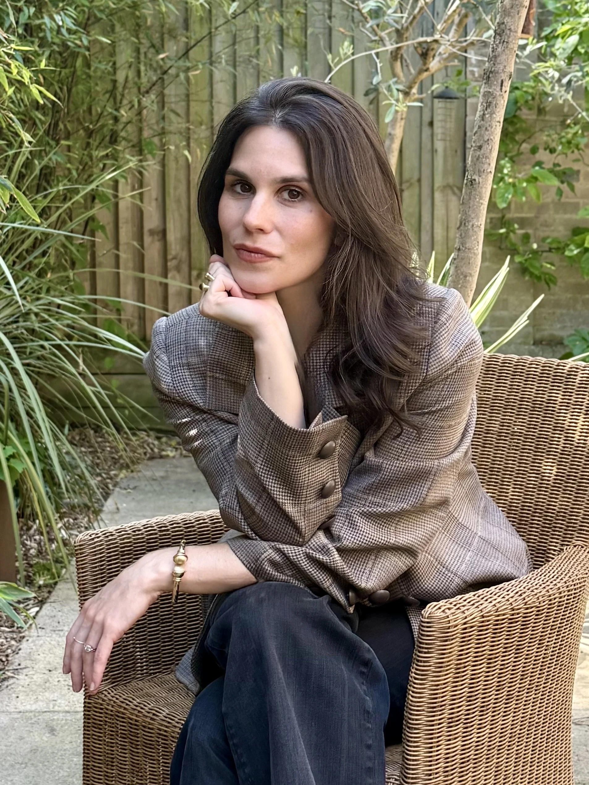 A woman with long brown hair sitting outdoors on a wicker chair, resting her chin on her hand, surrounded by green plants and trees in a garden setting.