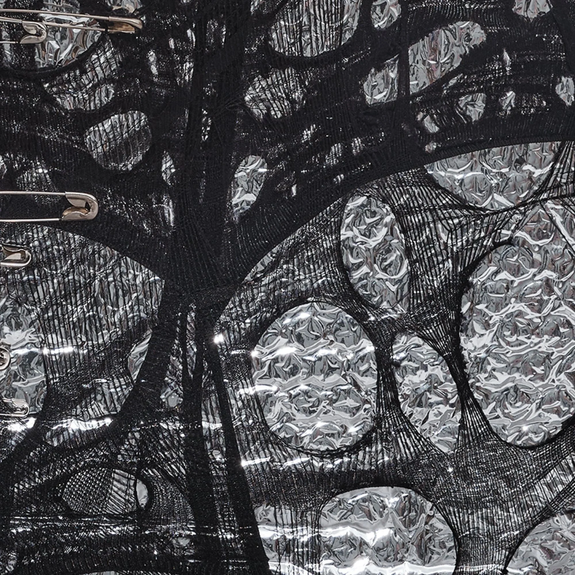 Textural detail of "Silver Dreams: Act 2 - Infinity" by Nicole Bajger, highlighting the interplay between the matte black synthetic textile and the reflective, metallic surface of the industrial packaging.