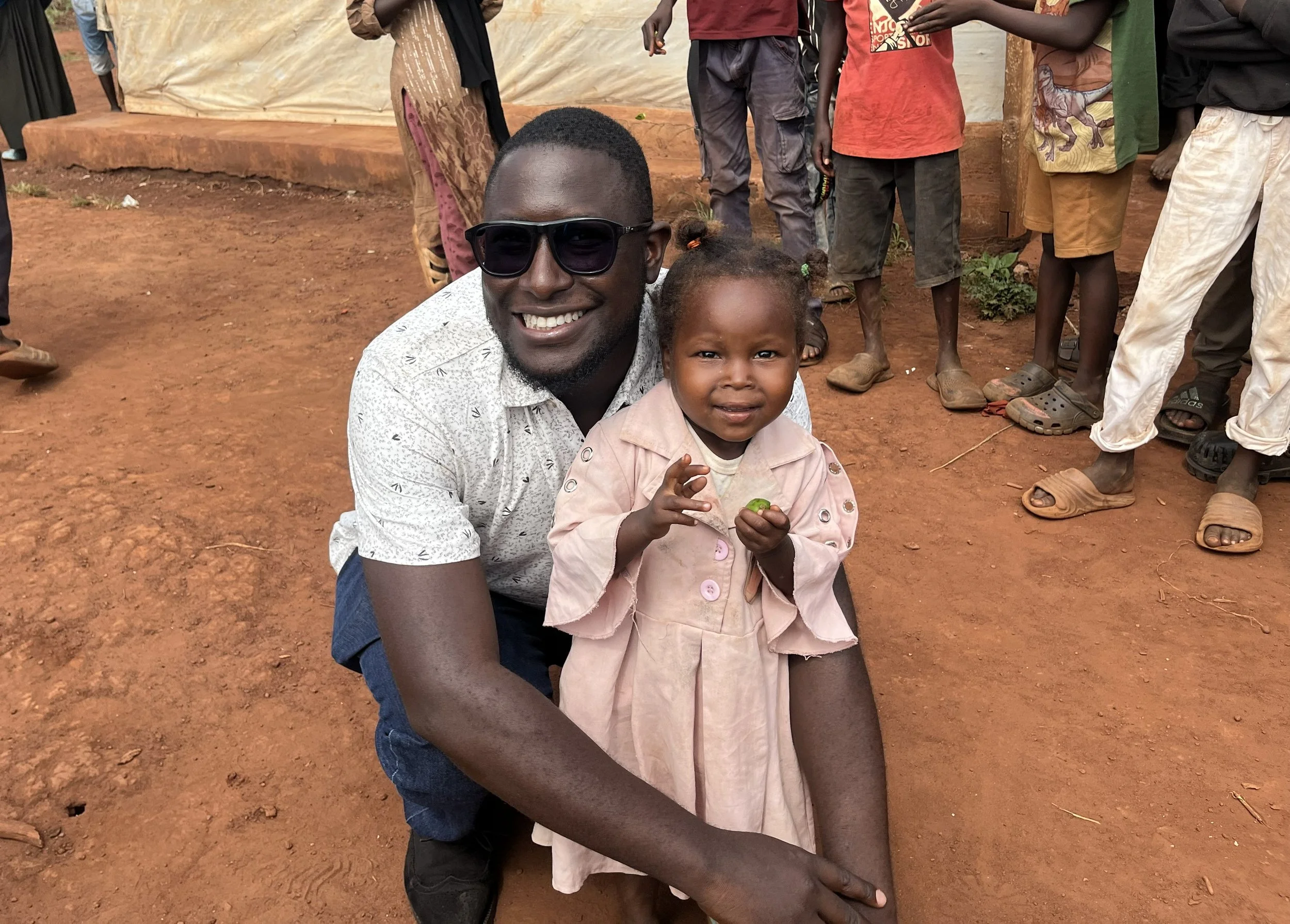An elderly man with a white beard wearing a dark shirt sits on the ground, holding a young girl with brown hair and a light green top. They are outdoors in front of a building with Arabic writing and a sign of a woman figure. The girl is smiling and looking up while the man is smiling at the camera.