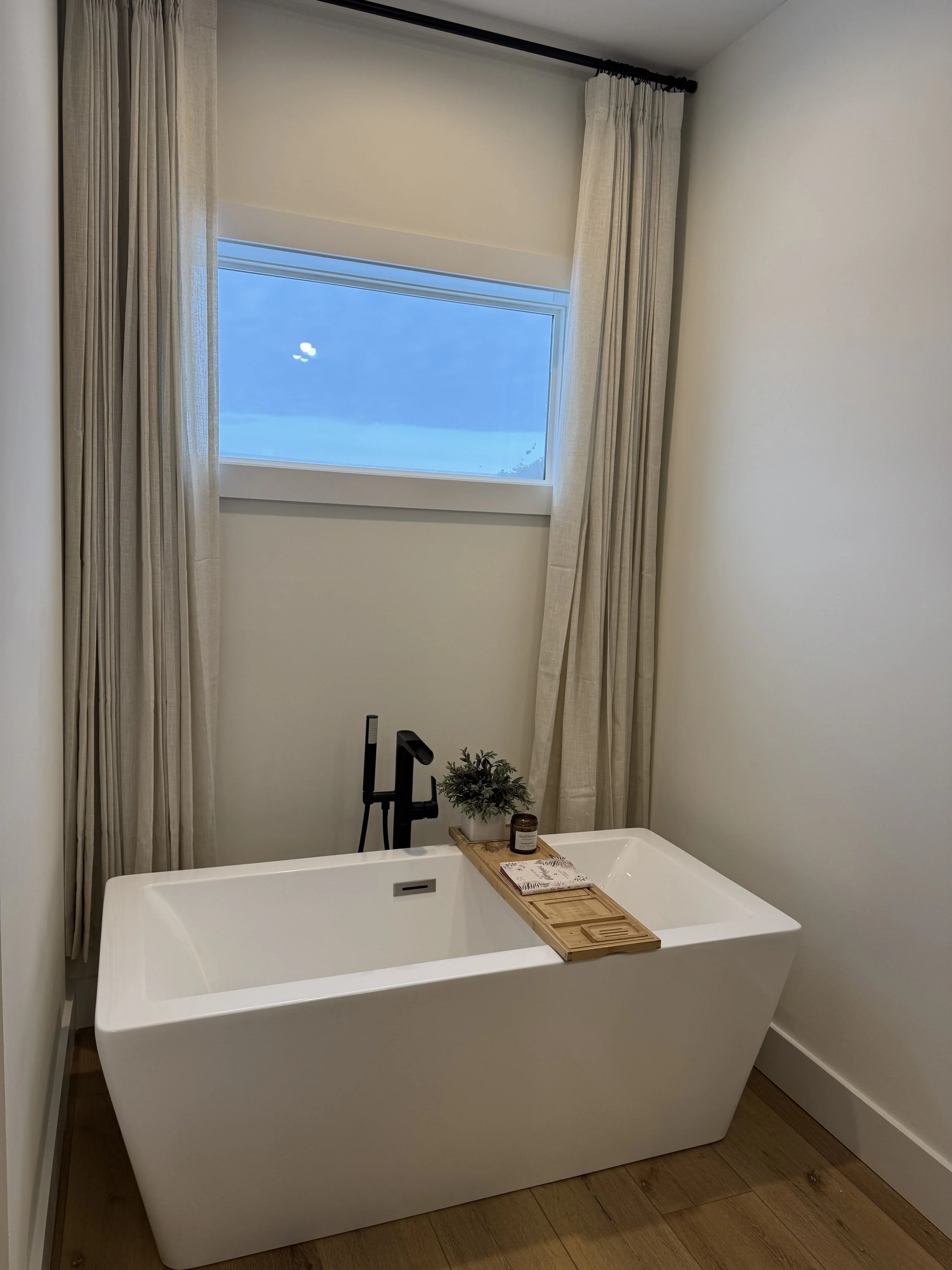 Modern bathroom with a white freestanding bathtub, a wooden bath tray holding a candle, a plant, and a book, beige curtains, a window showing a blue sky, and wooden floor.