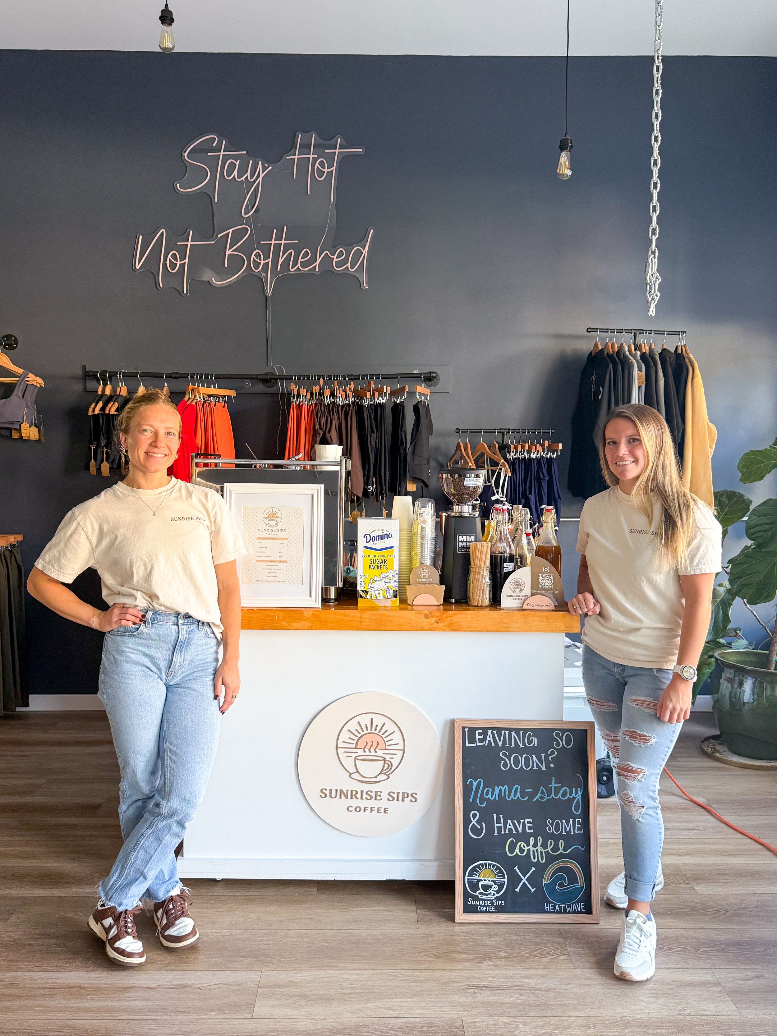 Two women standing in front of a coffee shop counter with a sign that reads "Sunrise Sips Coffee" and a chalkboard sign with a message about leaving soon, wearing casual clothes and smiling.