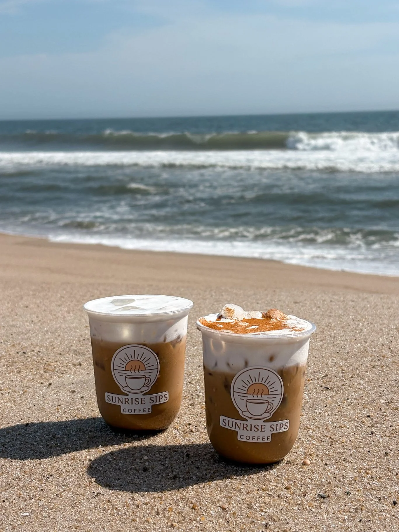 Two cups of coffee on the sand at the beach with the ocean and waves in the background.