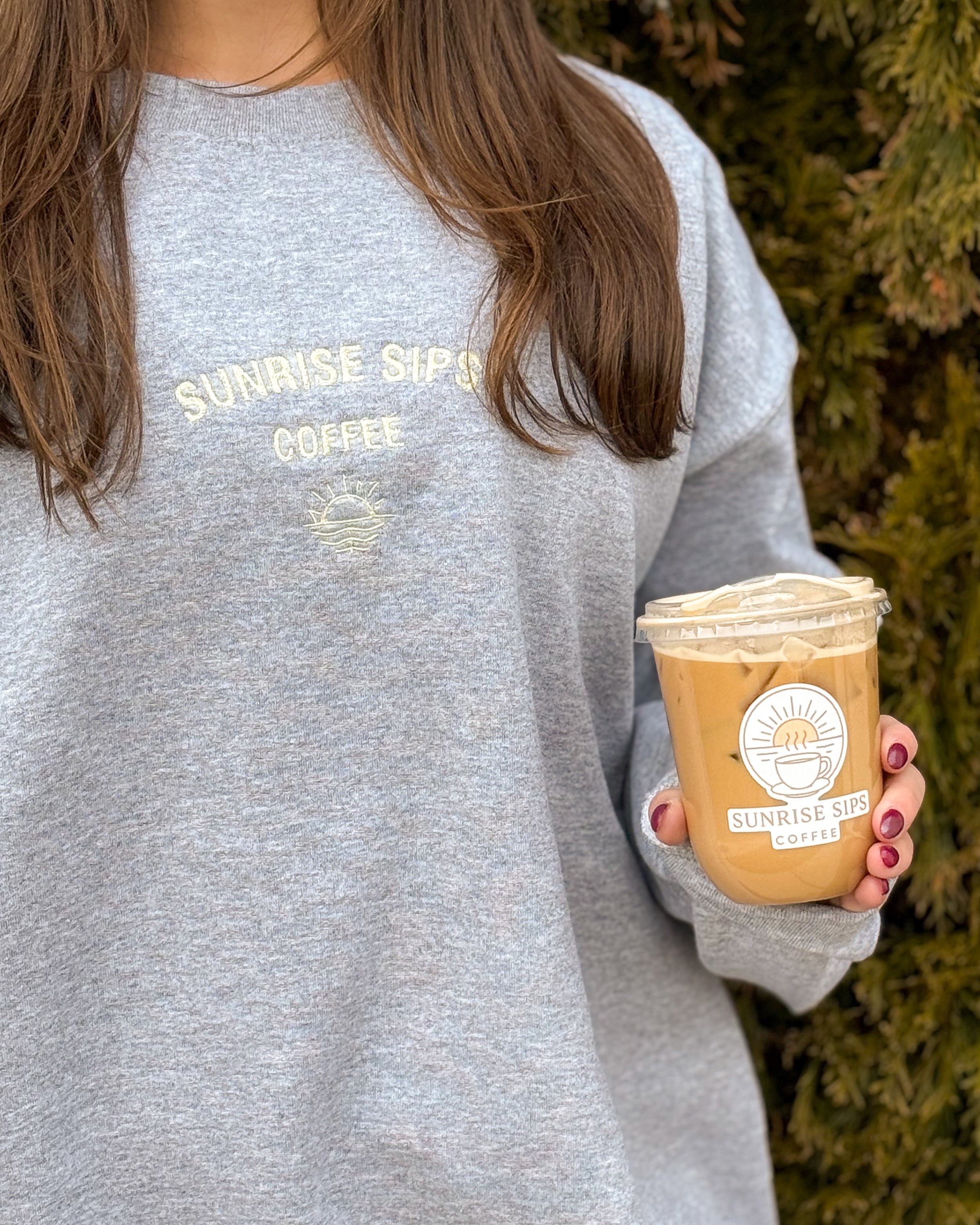Close-up of a woman in a gray sweatshirt holding a Starbucks coffee cup with a sunset logo and the words "Sunrise Sips" on it. She has long brown hair and is standing outdoors with greenery in the background.