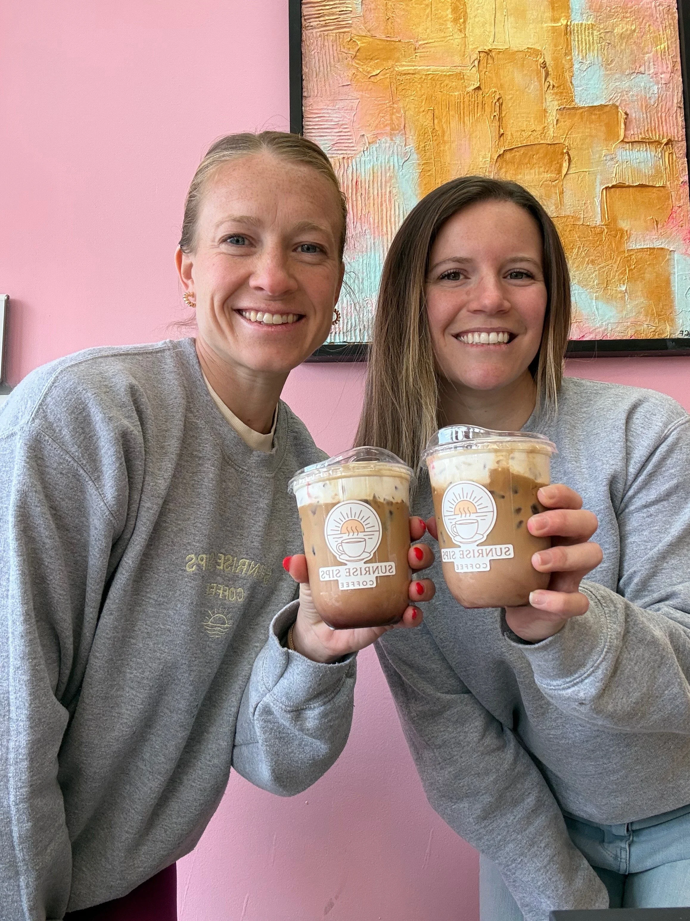 Two women smiling and holding cups of iced coffee in a cafe with pink walls and a colorful abstract art piece in the background.