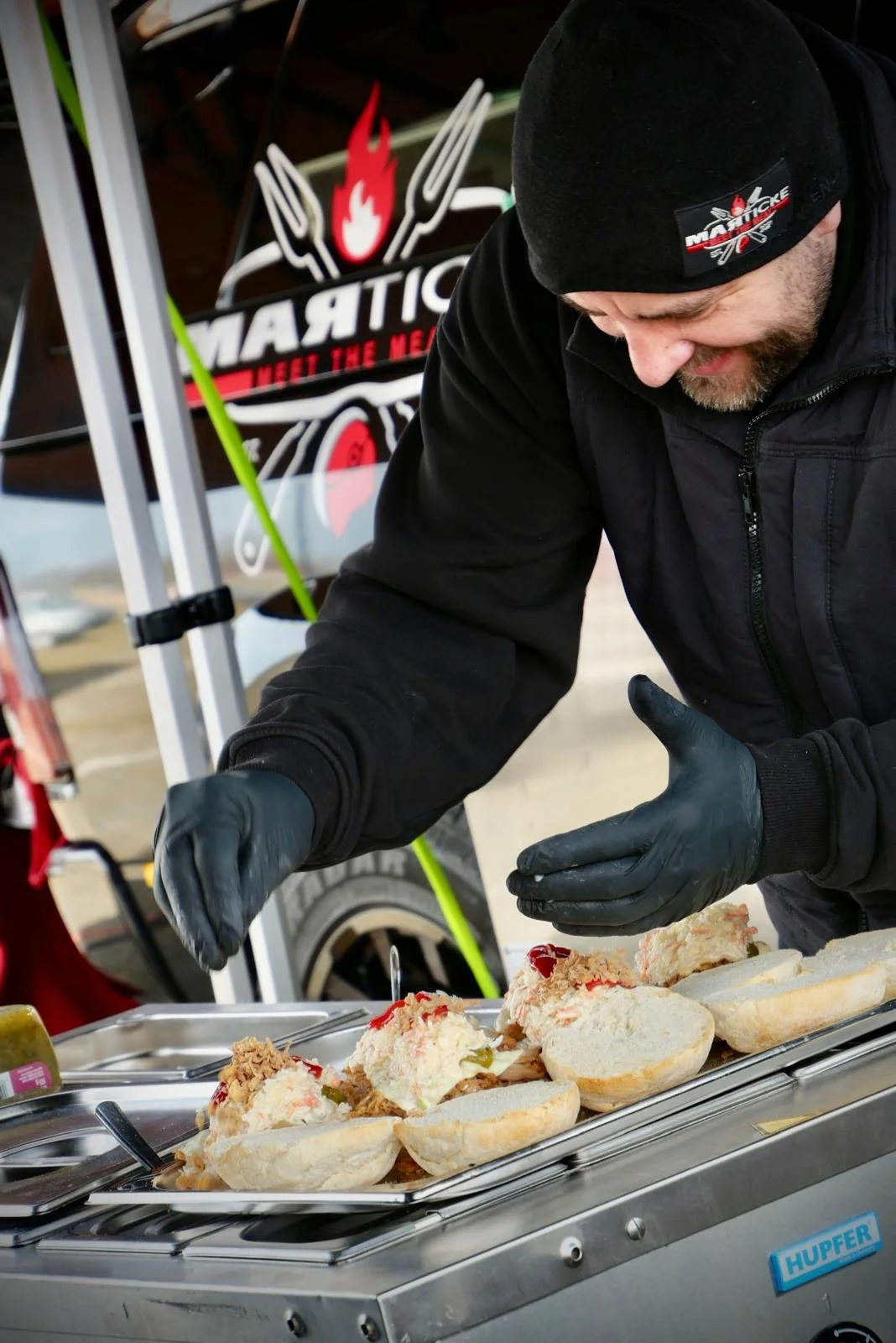 Ein Mann bereitet Sandwiches zu, mit einem Hintergrundbanner, das den Namen 'Martick' und die Slogans 'Meet the Meat' zeigt.