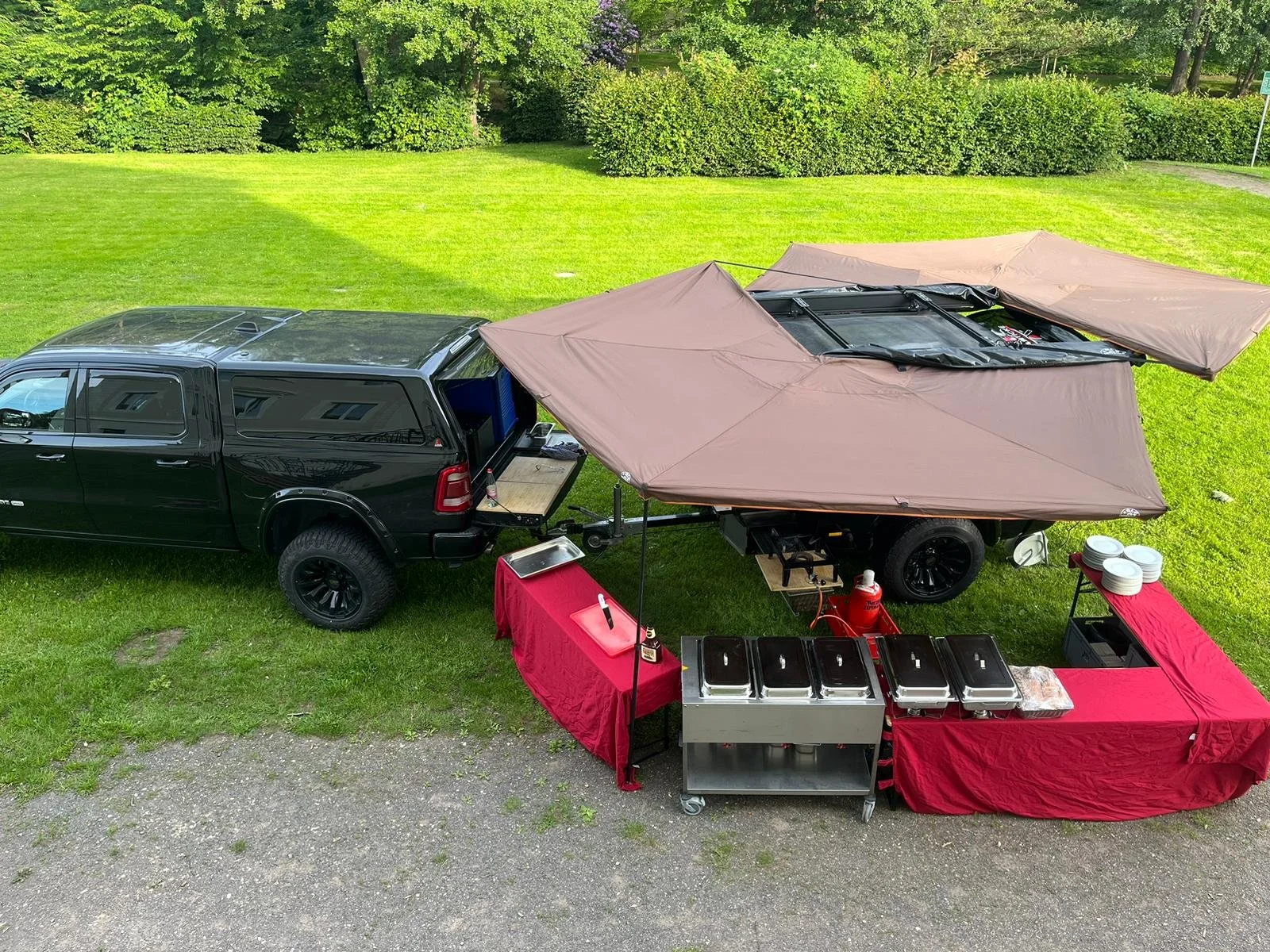 Ein schwarzer Pick-up-Truck mit Dachzelt, das auf der Ladefläche aufgebaut ist, neben einem Essensstand mit roter Tischdecke und mehreren chafing dishes im Freien auf einer grünen Wiese.