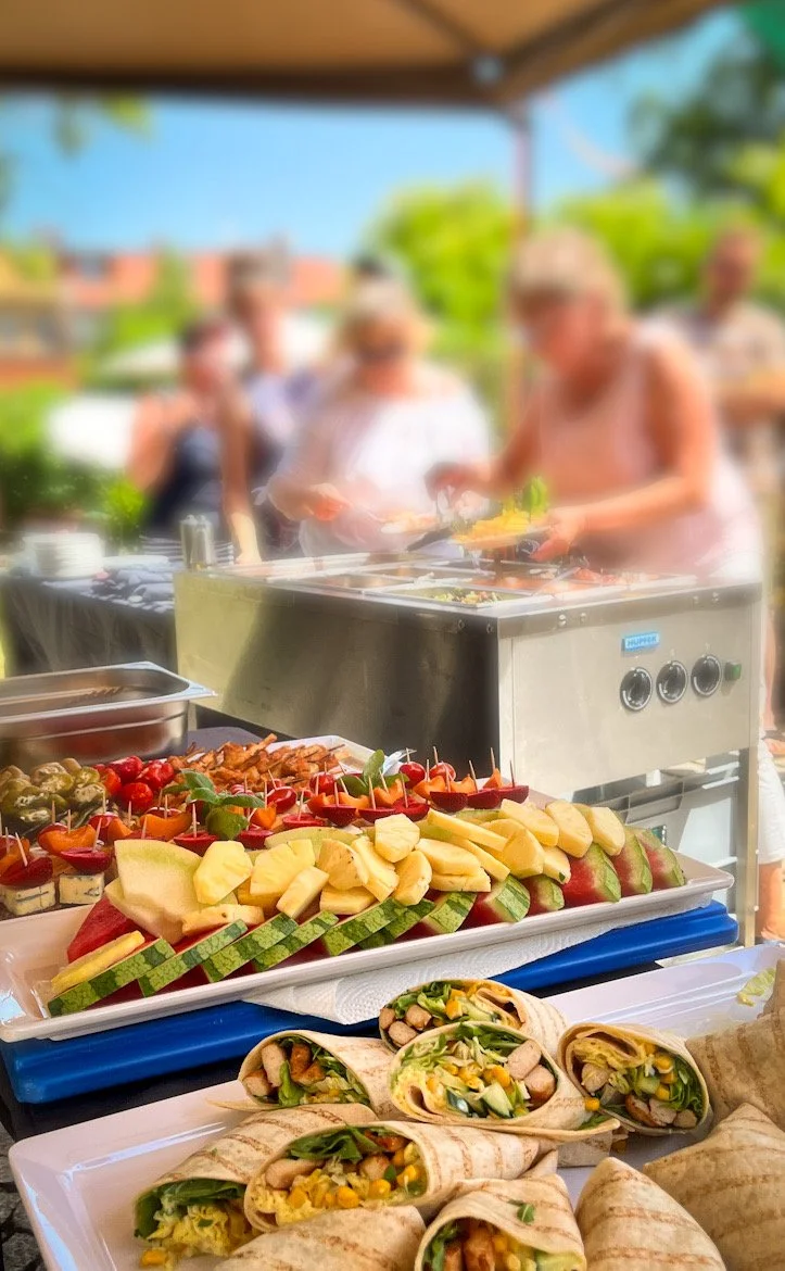 Buffet mit frischem Obst, Wraps, und einem Grill bei einer Gartenparty, im Hintergrund grillende Personen.