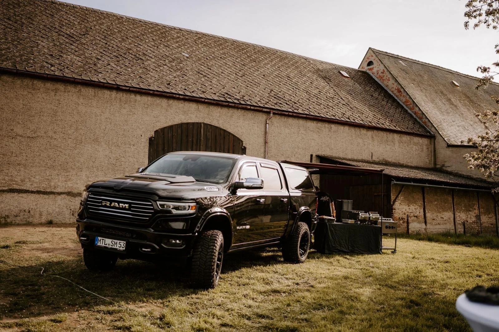 Schwarzer RAM Pickup-Truck steht auf einer Wiese vor einem alten Bauernhaus, neben einem Grill und einem Tisch mit Getränken.