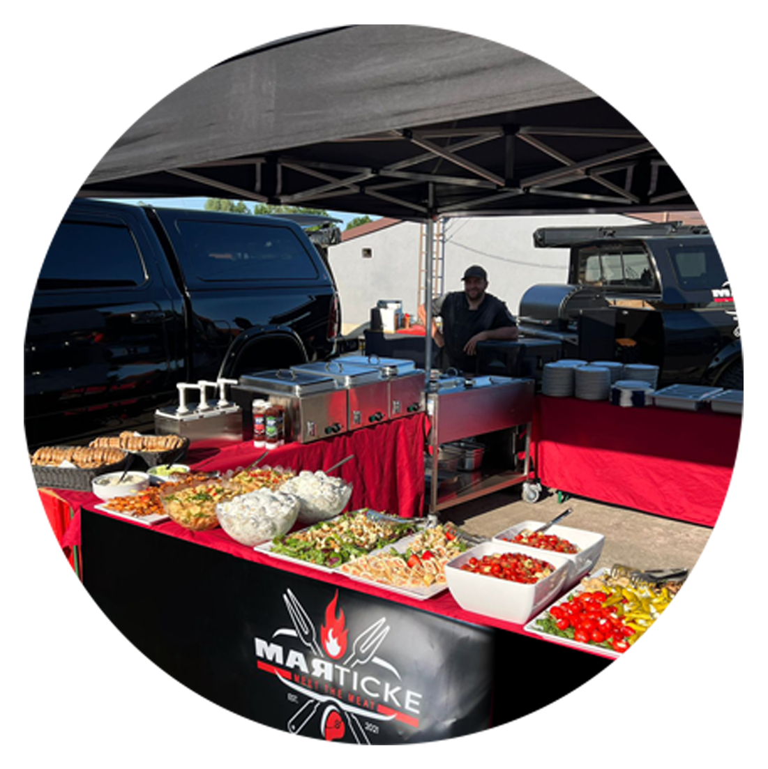 Stand mit Grillfleisch, Salaten und Beilagen bei einem Food-Event, mit einem Verkäufer im Hintergrund, unter einem Zelt, mit schwarzen Fahrzeugen im Hintergrund.