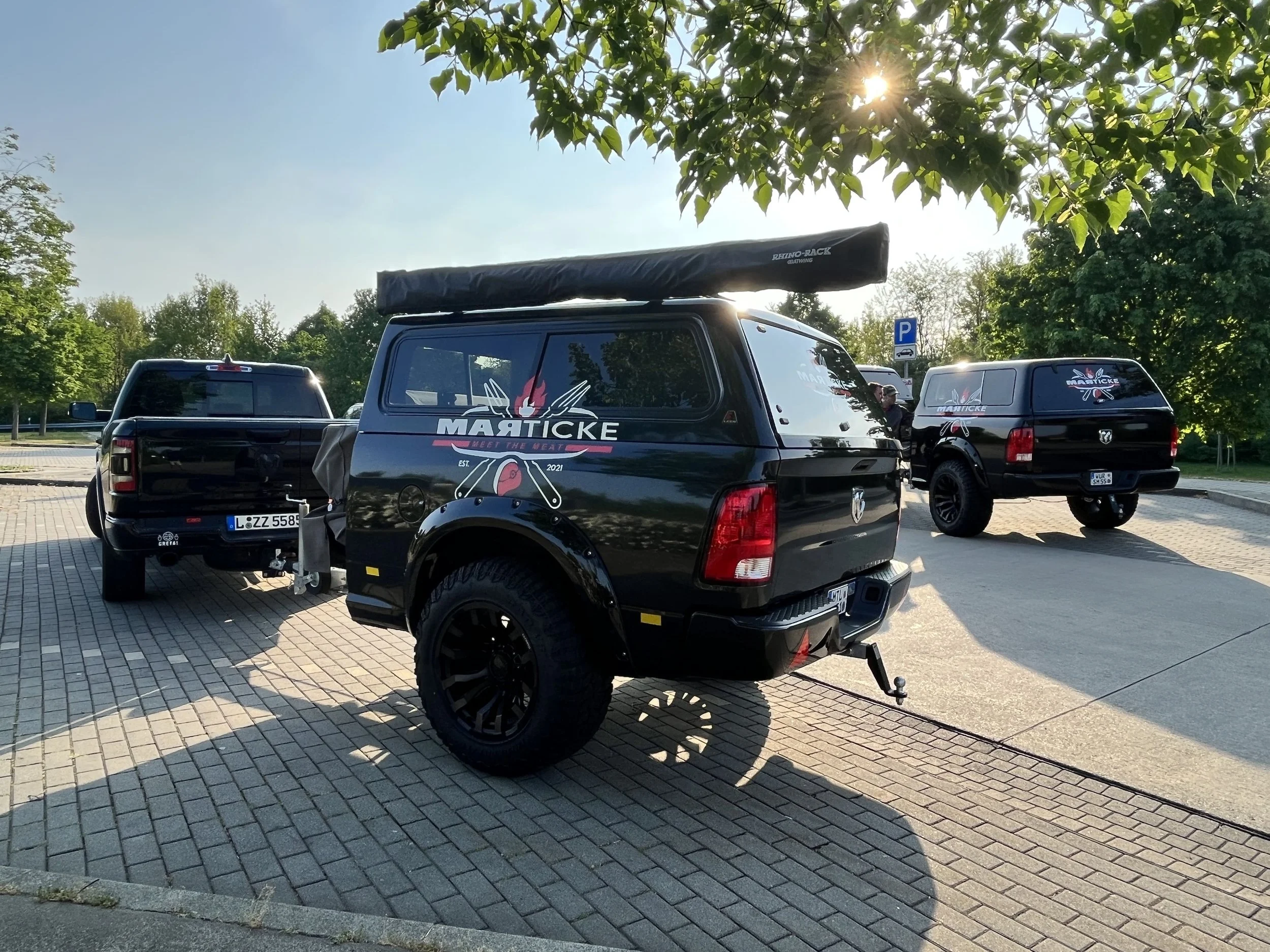 Parkplatz mit mehreren schwarzen Fahrzeugen, eines mit einer Markicke Dekoration und einem Dachzelt, unter Bäumen bei Sonnenlicht.