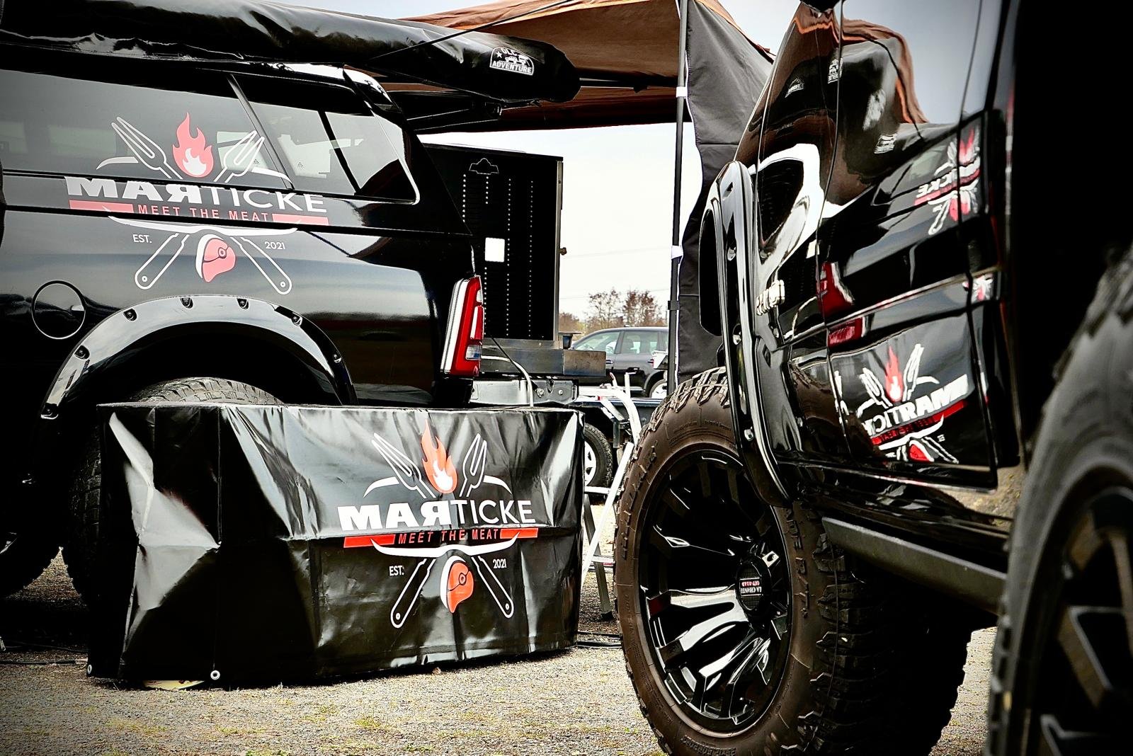 Verkaufsstand für Grillprodukte mit Black Trucks im Hintergrund, branding mit Messer, Gabel und Flamme, Marke 'MARTICKE', das Logo ist auf mehreren Gegenständen sichtbar.
