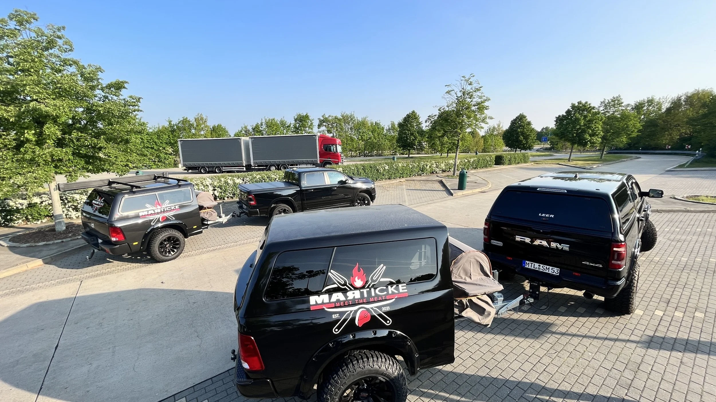 Mehrere Fahrzeuge, darunter Pickups und ein LKW, parken auf einem Parkplatz neben einer Straße mit Bäumen.