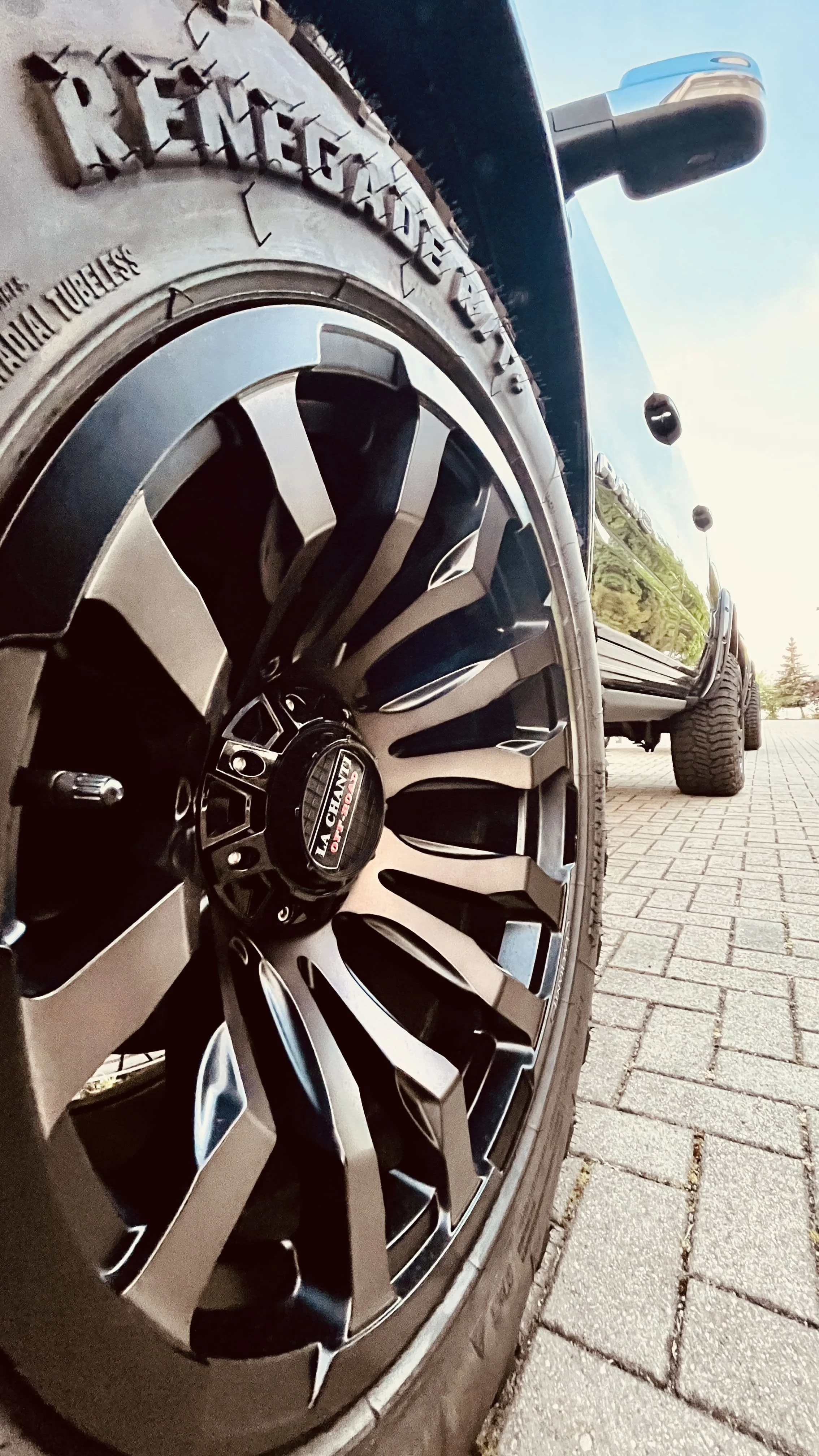 Nahaufnahme eines schwarzen Hyundai-Felgenrades mit Logo, Reifen auf einer gepflasterten Straße, im Hintergrund ein Teil eines schwarzen Autos und ein blauer Himmel.