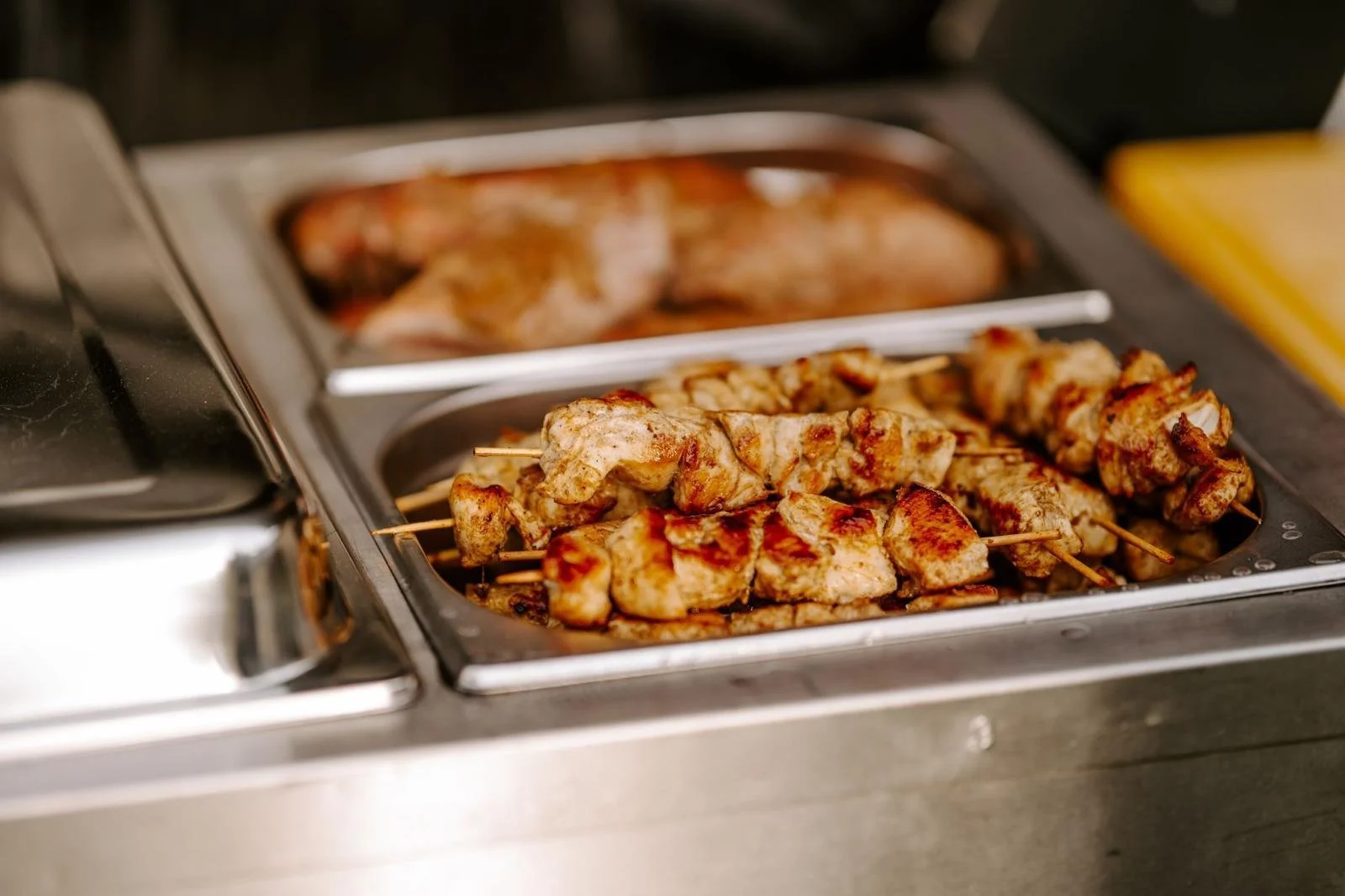 Hähnchenspieße liegen auf einer Wärmeschale in einem Buffet. Im Hintergrund ist eine weitere Speise, vermutlich Fisch oder Fleisch, sichtbar.