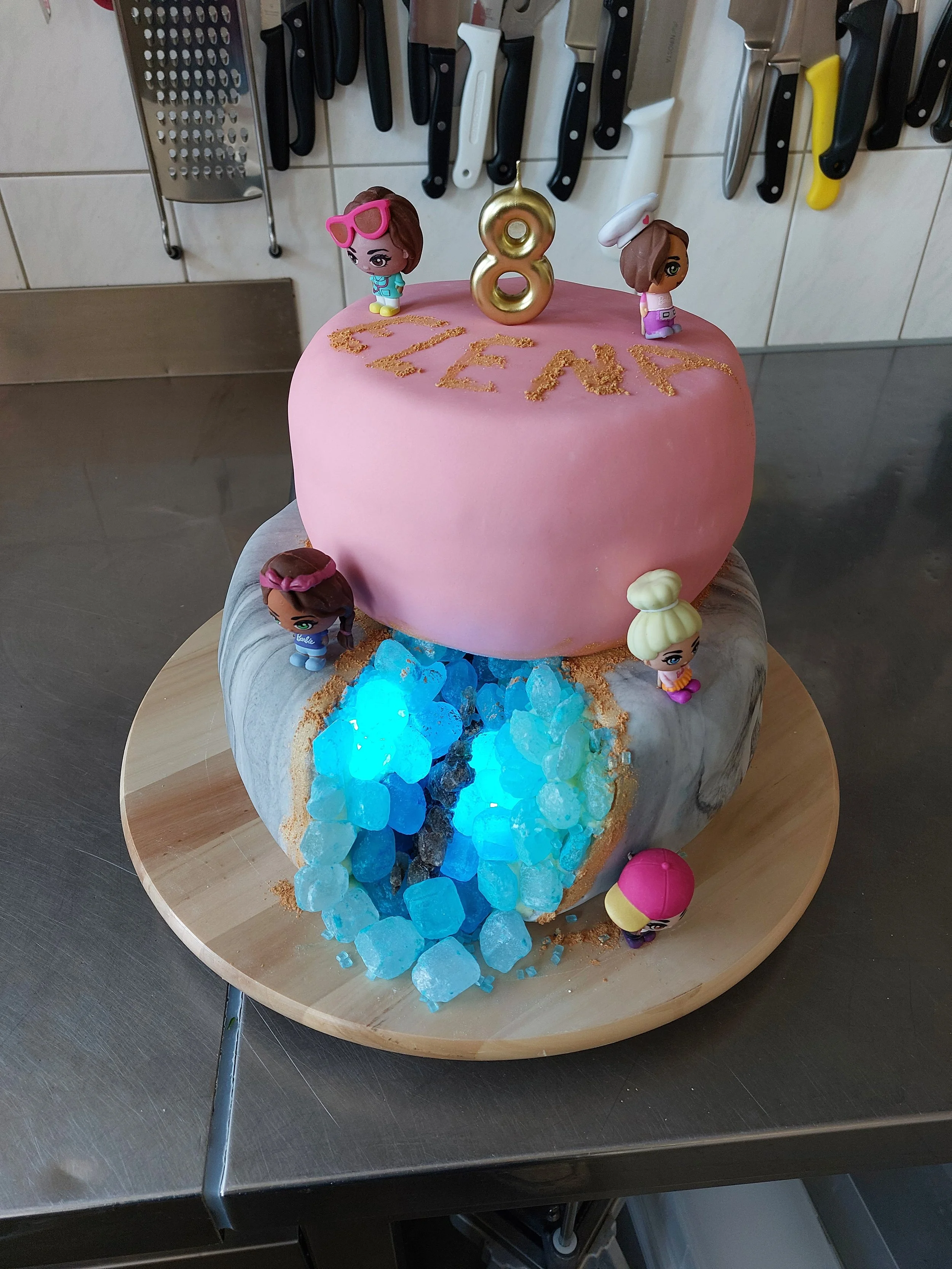 Eine Geburtstagstorte zum achtzehnten Geburtstag mit pinker Glasur, Kerzen in Form der Zahl 18, kleinen Barbie-Figuren und blauen dekorativen Steinen, die wie Wasser aussehen.