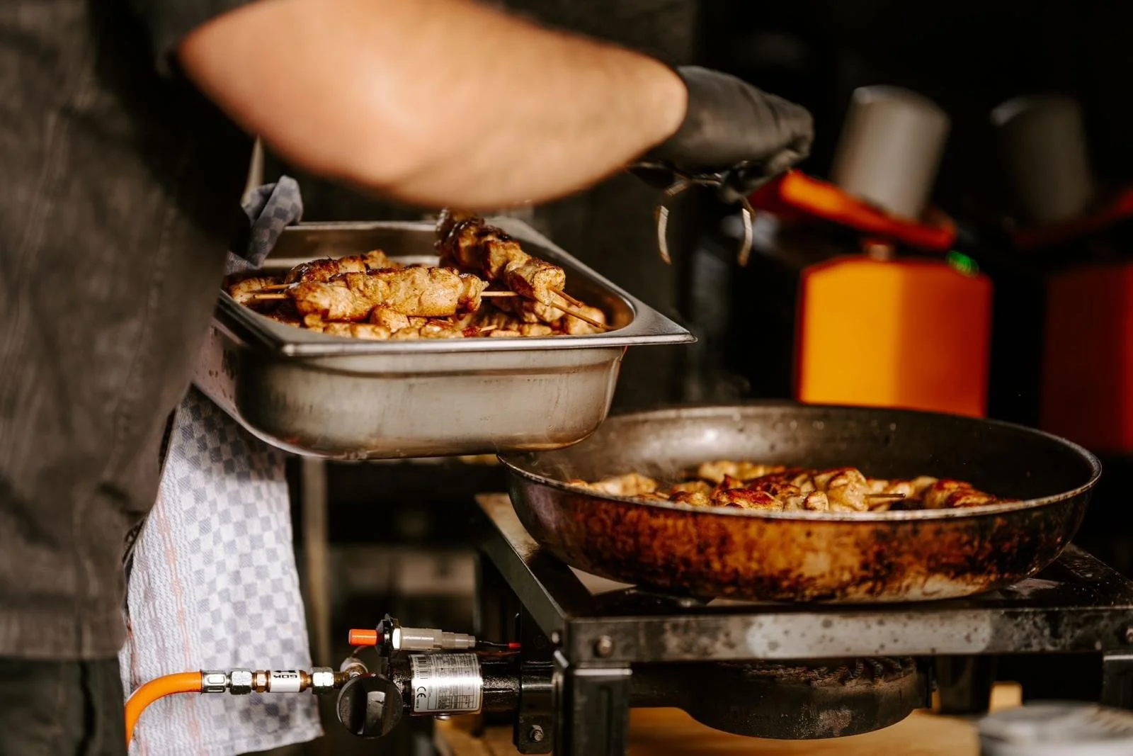 Person, die Grillspieße mit Fleisch auf einem Herd zubereitet, mit einem Emailblech voller fertiger Fleischspieße.