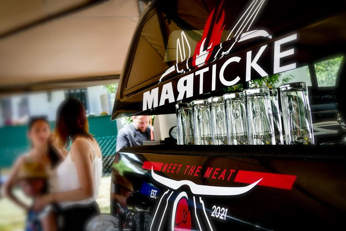 Eine Flipchart mit dem Logo und Slogan eines Food Trucks namens 'Martycke', der im Jahr 2021 gegründet wurde. Im Hintergrund sind Menschen beim Essen und Trinken zu sehen, vermutlich bei einem Festival oder einer Veranstaltung.