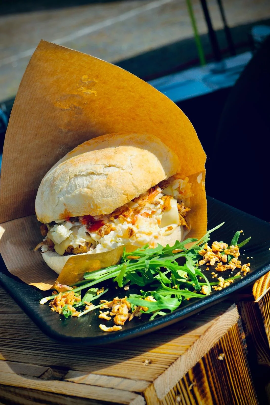 Sandwich mit Hühnerfleisch, Salat und Saucen auf einem schwarzen Teller, garniert mit Rucola und Croûtons, auf einem Holztablett.