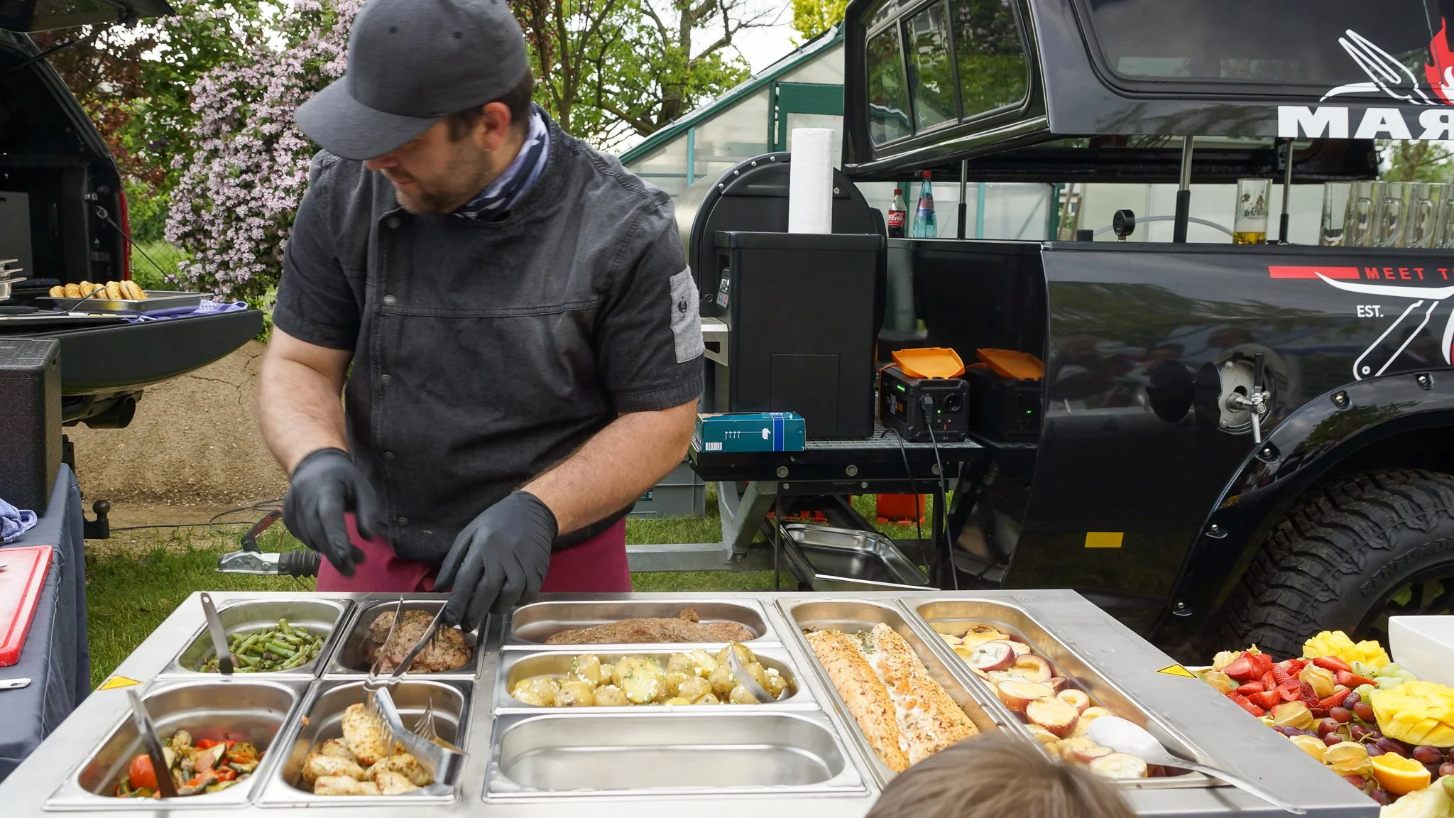 Mann beim Essen am Buffet mit verschiedenen warmen Speisen, auf einem mobilen Grillwagen im Freien.