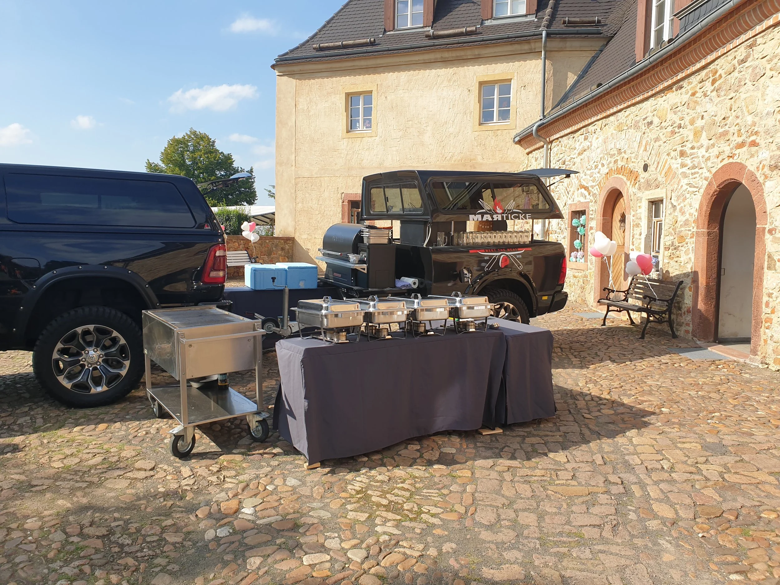 Ein Getränke- und Grillwagen steht in einem Hof vor einem Mehrfamilienhaus mit Steinmauer und Satteldach. Es sind Tische, Stühle, Ballons und Behälter in der Umgebung zu sehen.