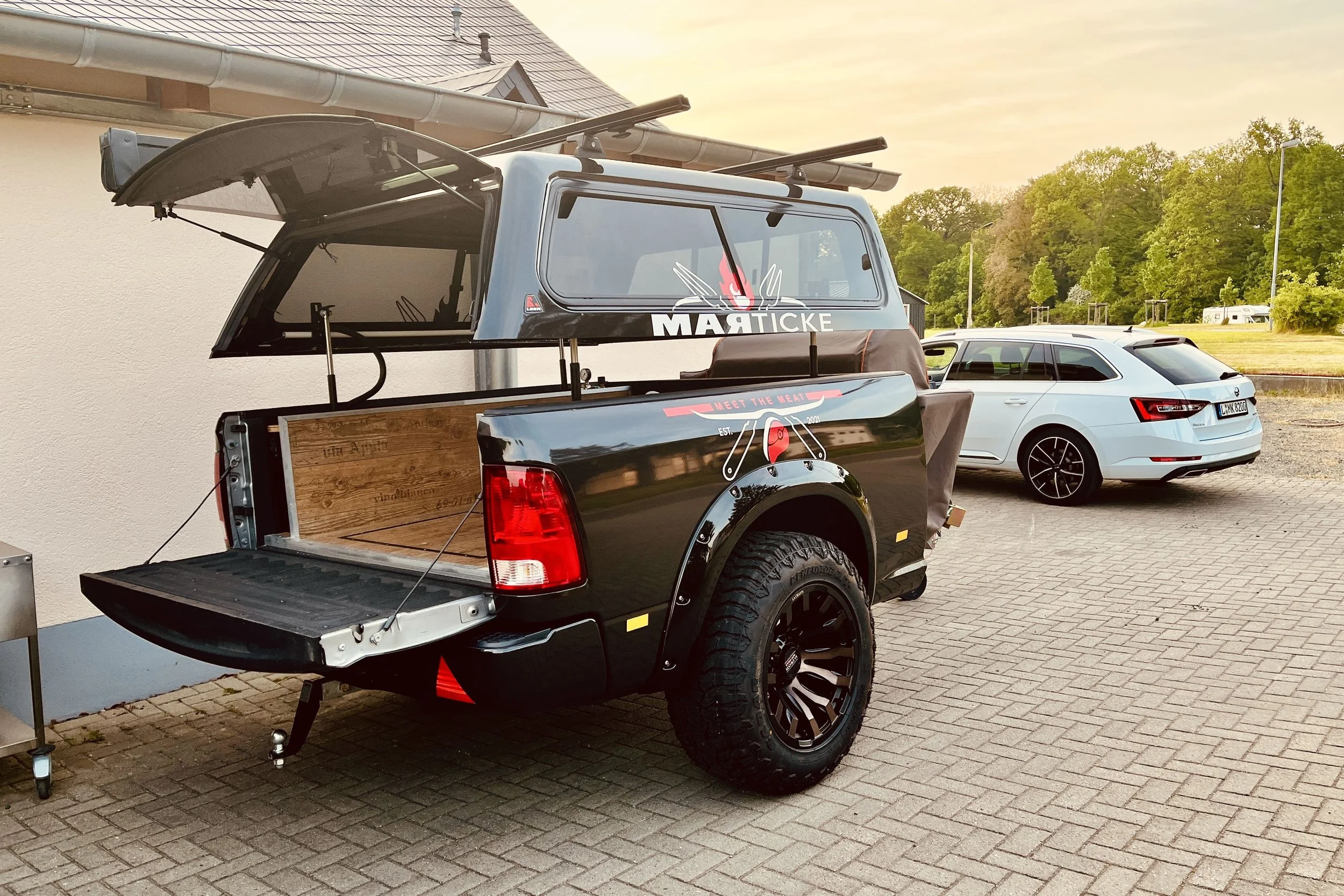 Ein schwarzer Pickup-Truck mit einem hölzernen Ladebett, das in der Nähe eines Gebäudes mit einer weißen Wand und einem Dach steht. Auf dem Truck sind Aufkleber mit dem Logo 'MARTICKE' und dem Slogan 'Meet the Heat'. Im Hintergrund sind grüne Bäume u