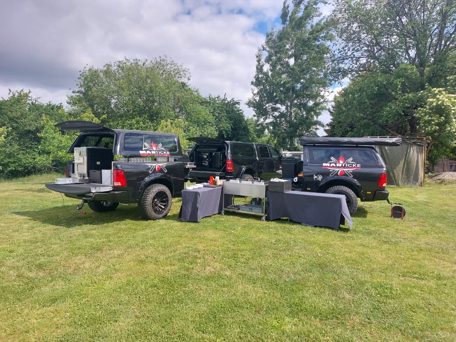 Zwei schwarze Pick-ups mit einem mobilen Küchen- und Grillbereich im Freien, auf einem Rasen, mit Bäumen im Hintergrund.