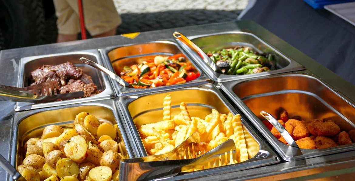 Verschiedene warme Speisen in einer Buffet-Station, darunter Kartoffeln, Pommes, Fleisch, Gemüse und Kroketten.