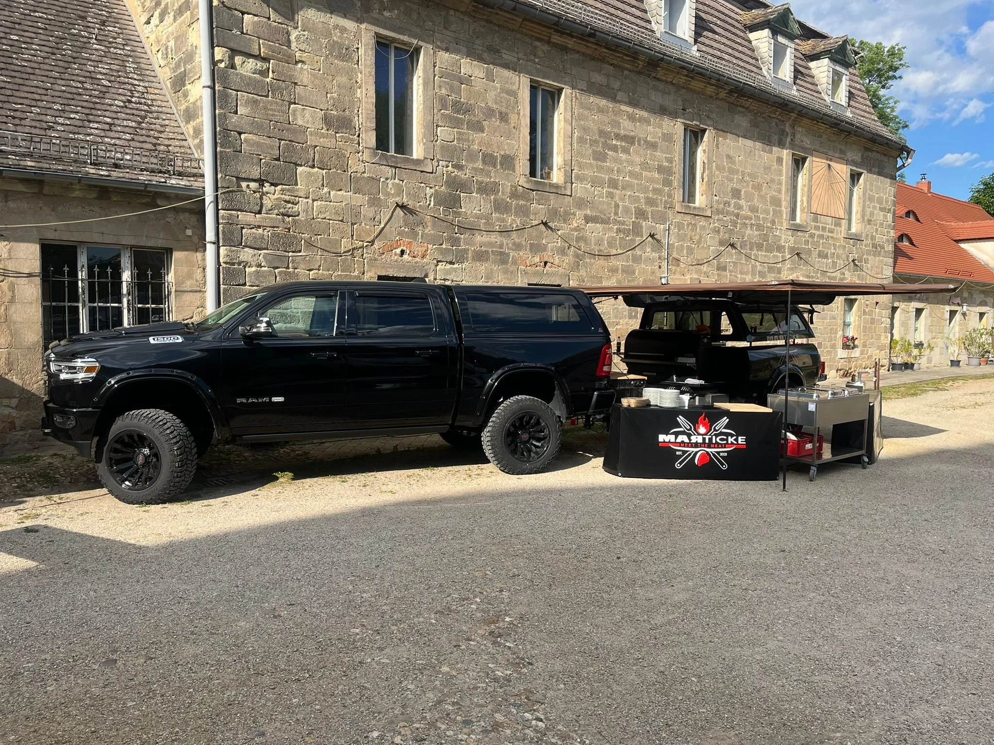 Ein altes Steinhaus mit mehreren Fenstern und einem roten Dach, davor stehen zwei schwarze Fahrzeuge, eines davon ist ein Pick-up-Truck mit einem Grill und einer Grillstation im Vordergrund.