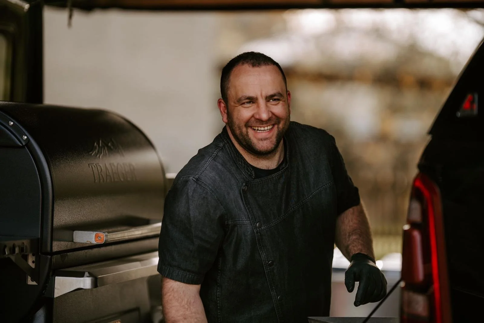 Ein lächelnder Mann in dunkler Jacke beim Grillen im Freien, neben einem schwarzen Grill und einem schwarzen Auto.