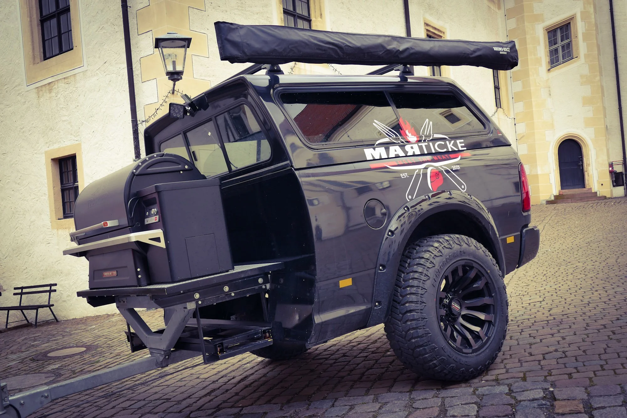 Ein schwarzer Offroad-Van mit einem Logo und Text auf der Seite, angehängt an ein Fahrzeug, das im Freien auf Kopfsteinpflaster steht, vor einer gelben Gebäudefassade.