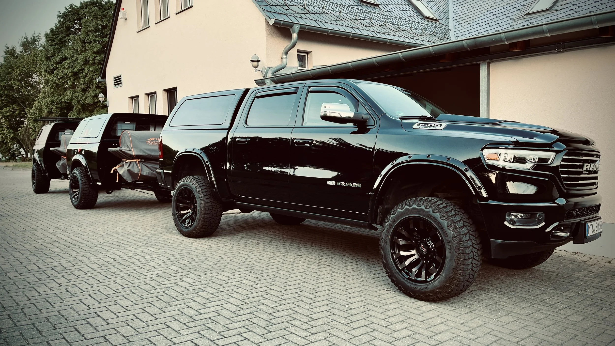 Schwarzer Pick-up Truck mit aufgemachten Rädern und ein Heckzelt, parkend auf einer gepflasterten Fläche vor einem Gebäude mit beige Wänden und grauem Dach.