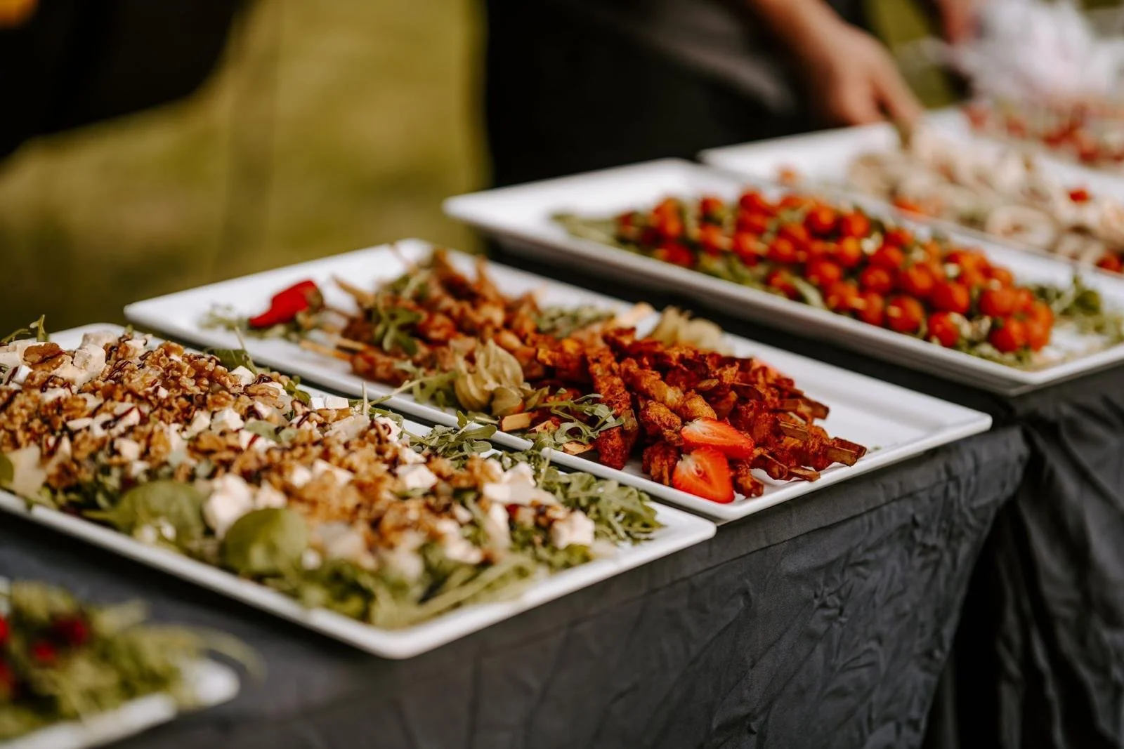 Verschiedene geordnete Salate und Vorspeisen auf weißen rechteckigen Tellern, auf einem schwarzen Tuch bedeckt. Im Hintergrund sind Menschen. Das Essen enthält Tomaten, Fleisch, Käse und grüne Blätter.