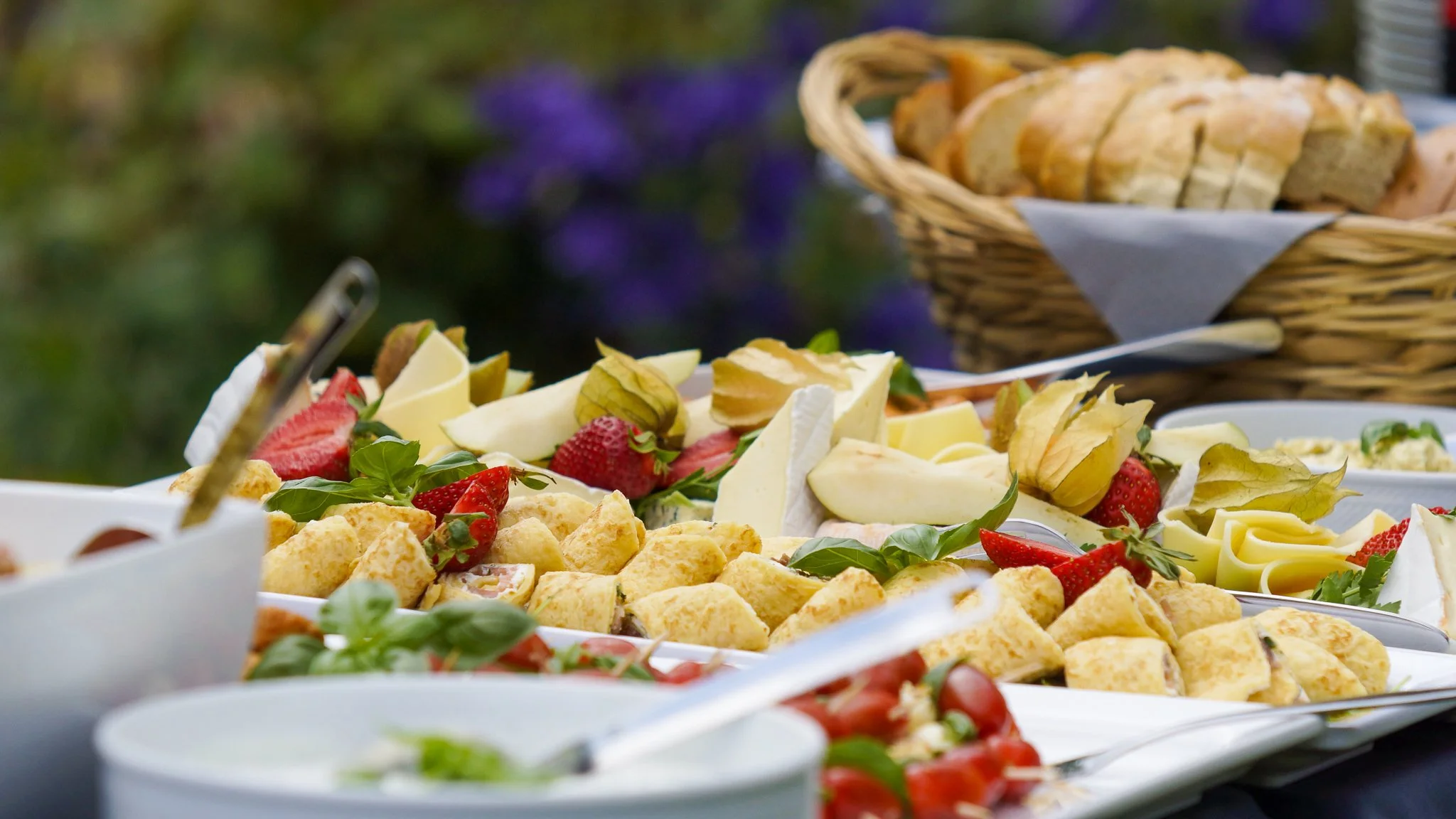 Verschiedene Speisen und Snacks auf einem Buffet, darunter Erdbeeren, Käse, Wurst, Brot und andere Häppchen, bei einer Gartenparty.