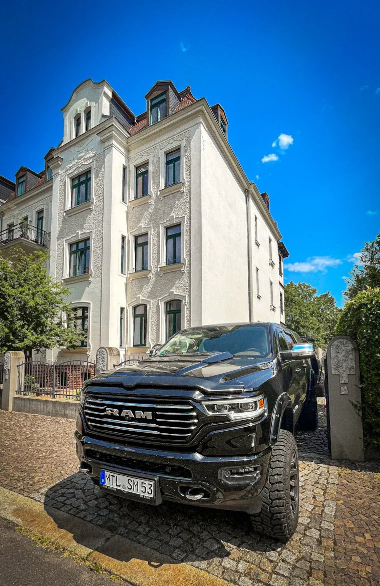 Ein schwarzer RAM Pickup vor einem weißen mehrstöckigen Wohngebäude mit blauen Fensterrahmen, grünem Baum und einem blauen Himmel mit Wolken.