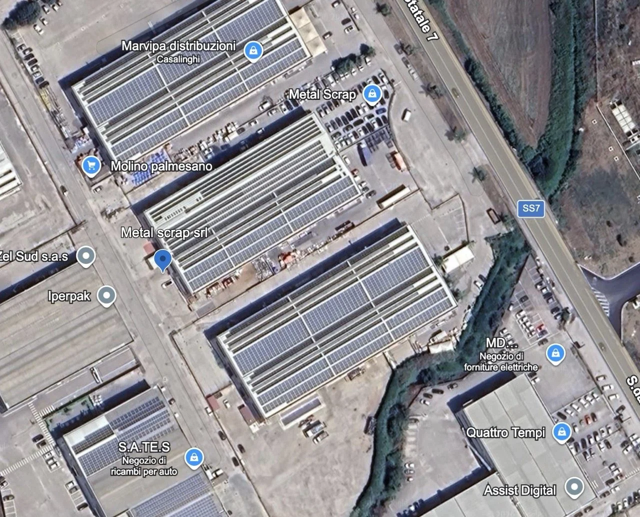 Immagine satellitare di una zona industriale con edifici industriali dotati di pannelli solari sui tetti, parcheggi e strade, inclusa la strada SS7 e vari punti di interesse come negozi di ricambi auto, distributori di materiali di scarto e un negozio di forniture elettriche.