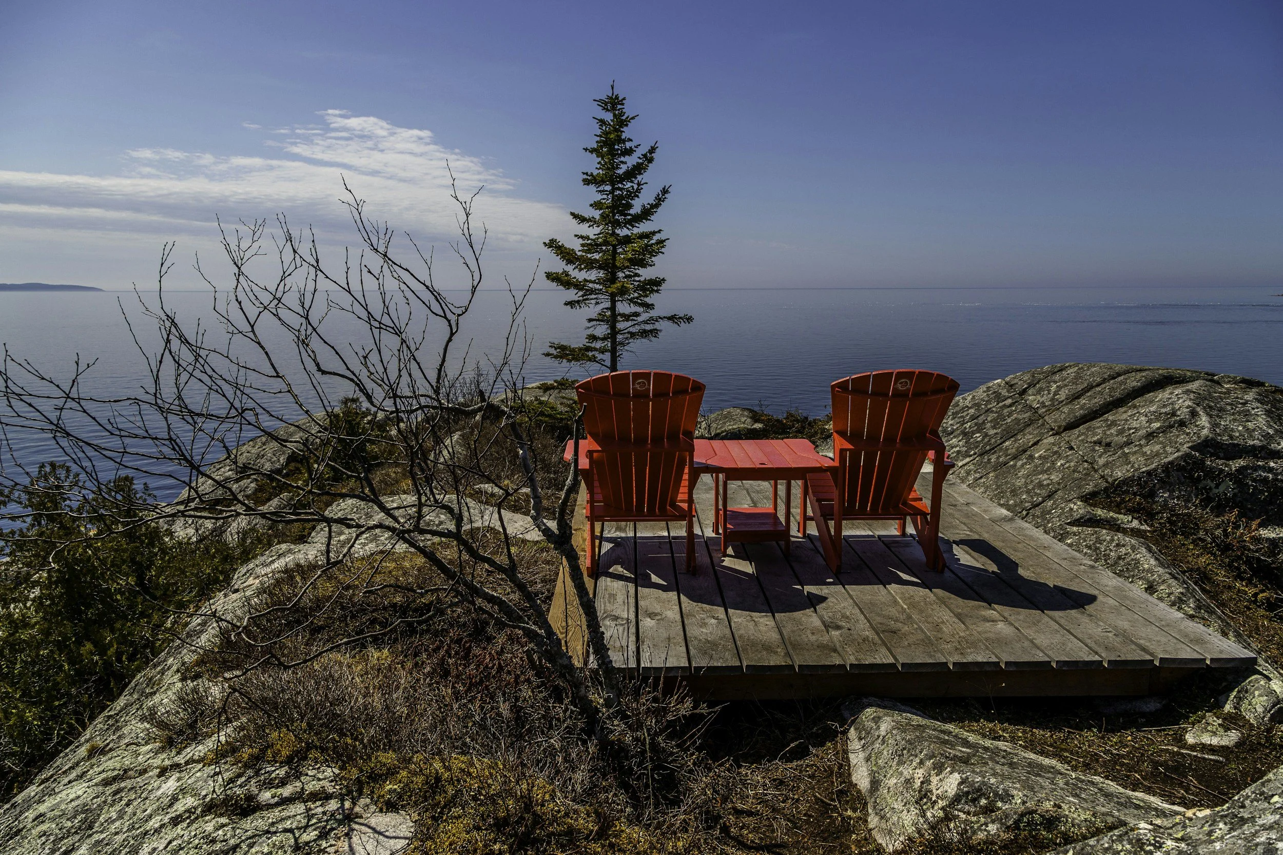 Two red Adirondack chairs on a wooden deck overlooking a calm body of water with a small pine tree nearby and a mostly clear sky.