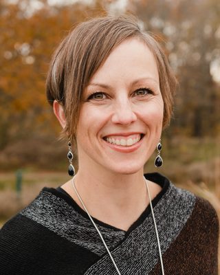 A woman with short light brown hair and earrings smiling outdoors with autumn-colored trees in the background.
