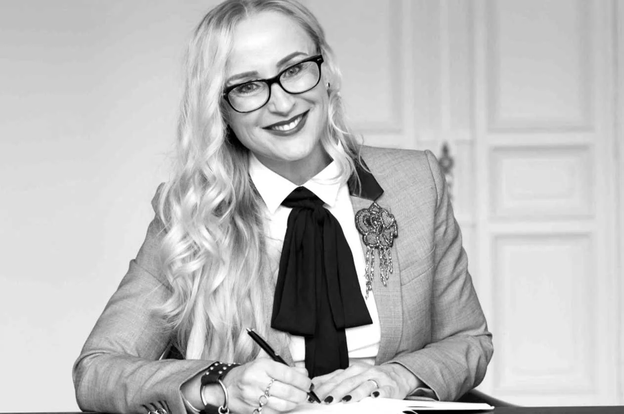 A woman with long wavy blonde hair, wearing glasses, a gray suit, a white shirt, and a black bow tie, is smiling and holding a pen at a desk.