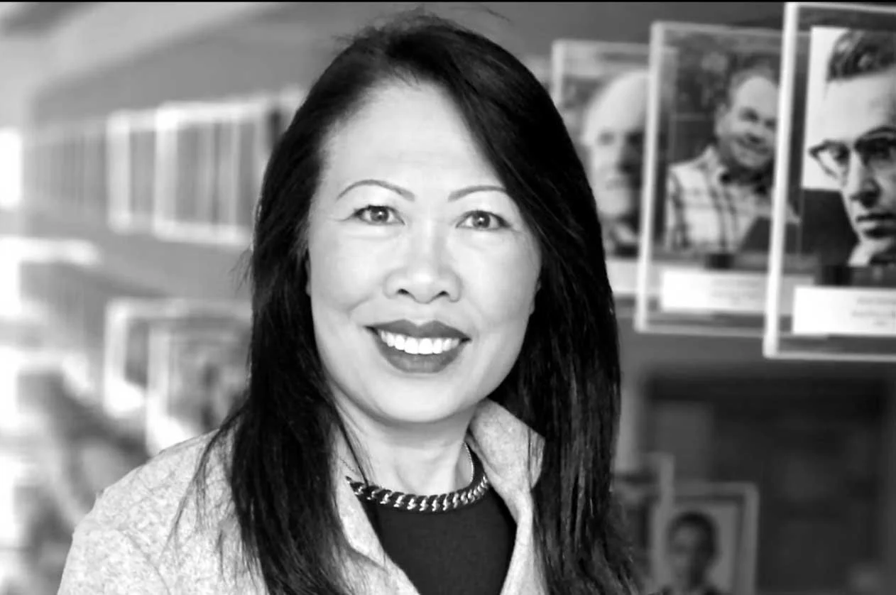 Black and white photo of an Asian woman with long dark hair smiling, standing in a room with shelves of books and photos behind her.